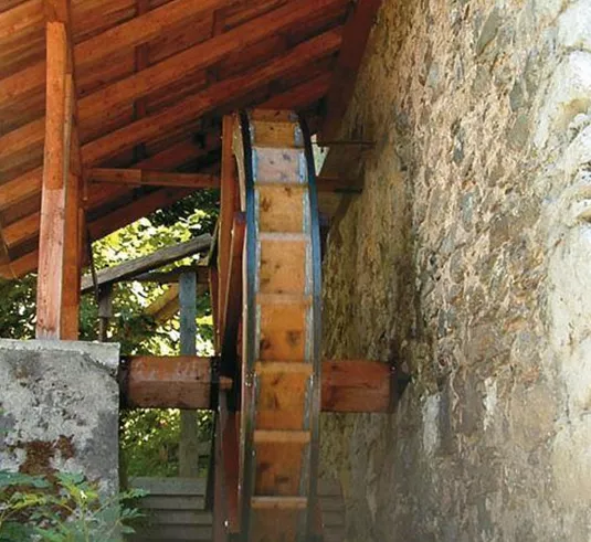 Ein Wasserrad aus Holz steht an einer steinernen Wand. Es ist teilweise durch ein Dach geschützt und von grünen Pflanzen umgeben.