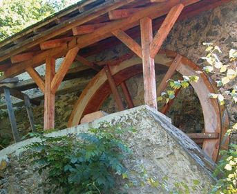 Ein Wasserrad aus Holz, das unter einem überdachten Bau steht. Umgeben von Pflanzen und einem klaren Himmel.