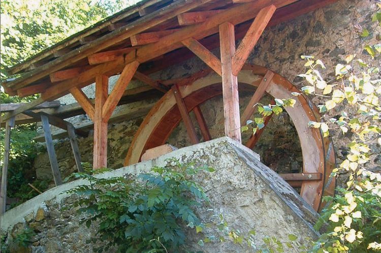 Ein Wasserrad aus Holz, das unter einem überdachten Bau steht. Umgeben von Pflanzen und einem klaren Himmel.