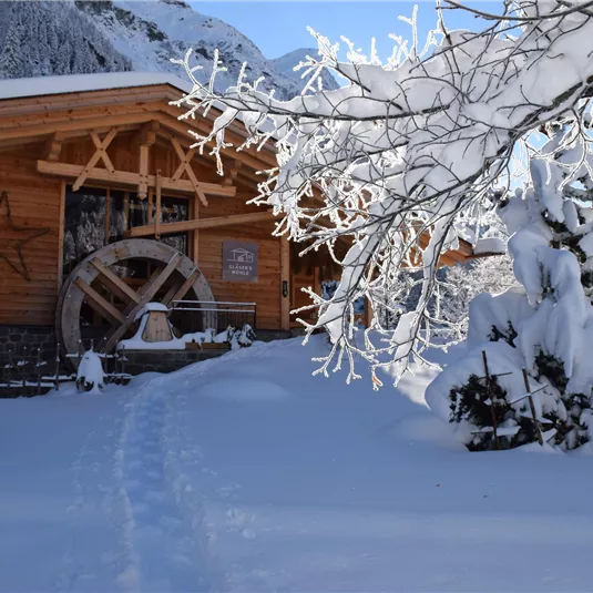 Ein gemütliches Holzhaus im Schnee mit einem großen Wasserrad. Die Umgebung ist winterlich und friedlich, mit schneebedeckten Bäumen.