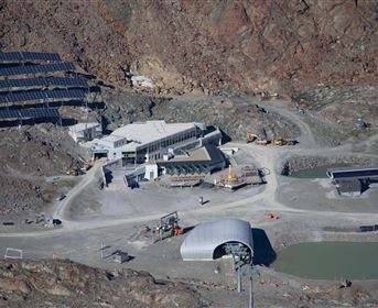Eine moderne Anlage in einer gebirgigen Landschaft, umgeben von Solarpanelen. Im Vordergrund sind mehrere Gebäude und ein kleiner Teich zu sehen.