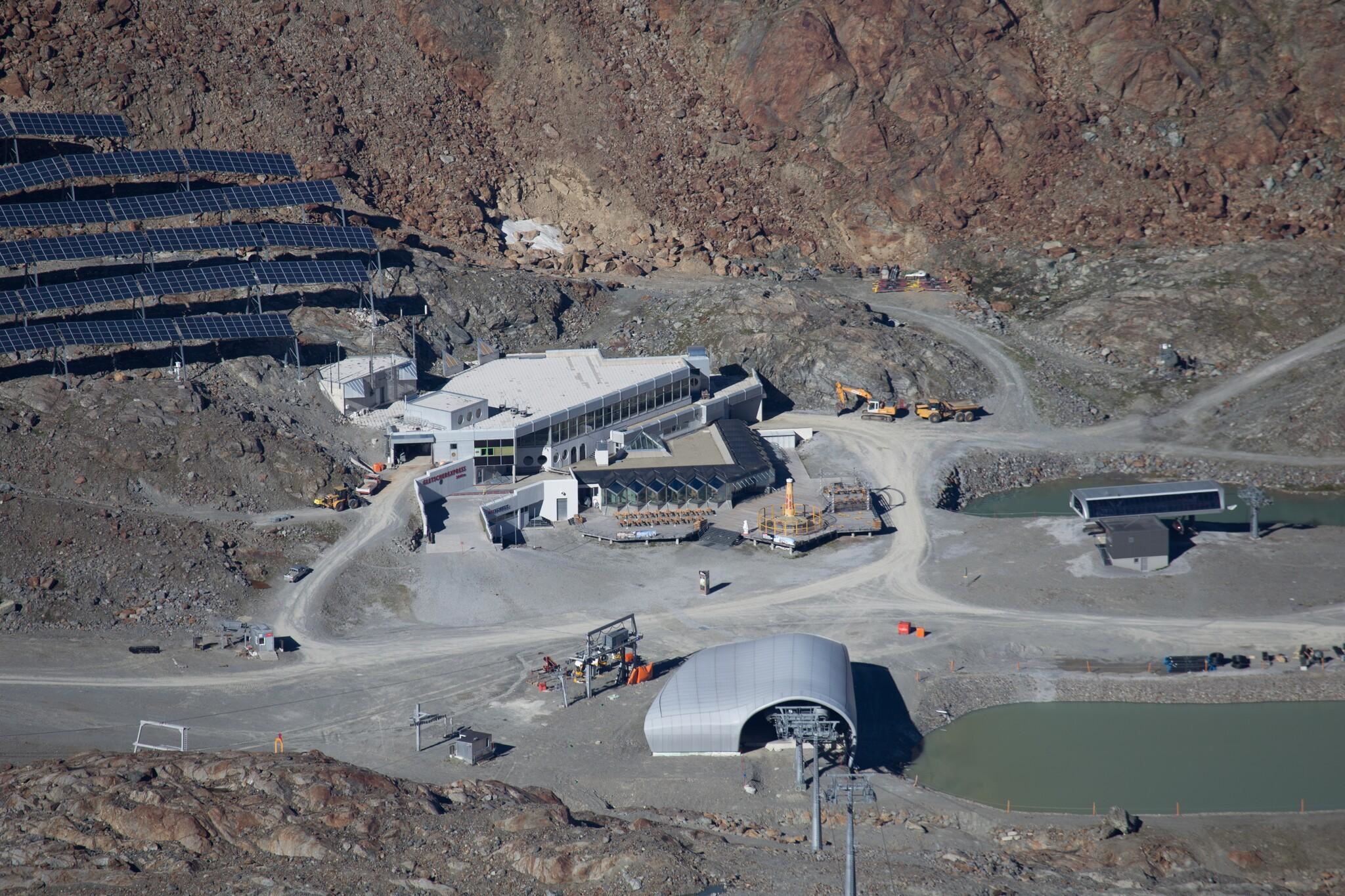 Eine moderne Anlage mit Solarpanelen in einer bergigen Umgebung. Es gibt verschiedene Gebäude und einen Wasserbereich in der Nähe.
