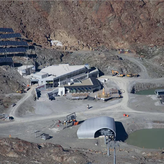Eine moderne Anlage mit Solarpanelen in einer bergigen Umgebung. Es gibt verschiedene Gebäude und einen Wasserbereich in der Nähe.