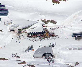 A bustling ski resort in the snow with numerous people. In the background, mountain peaks and solar panels can be seen.