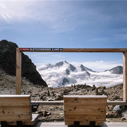 A wooden frame with a view of snow-covered mountains and a clear sky. In the foreground, there are wooden benches for viewing the landscape.