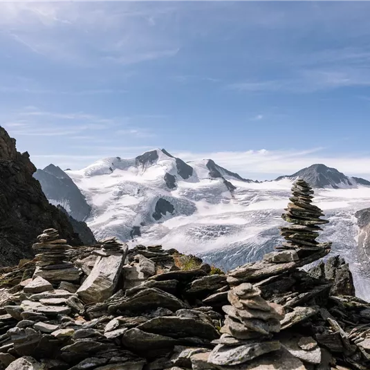 An impressive mountain landscape with snow-covered peaks and a clear blue sky. In the foreground, there are some stone cairns on a rocky surface.