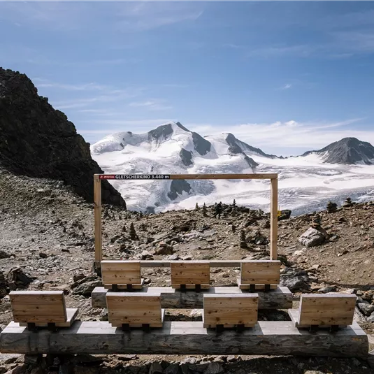 A stable viewpoint with a wooden frame in front of majestic mountains and glaciers. The clear sky and the impressive landscape invite contemplation.