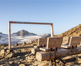 Ein rustikaler Ausblicksrahmen auf einem Berggipfel. Im Hintergrund sind Gipfel und Gletscher in der Ferne sichtbar.