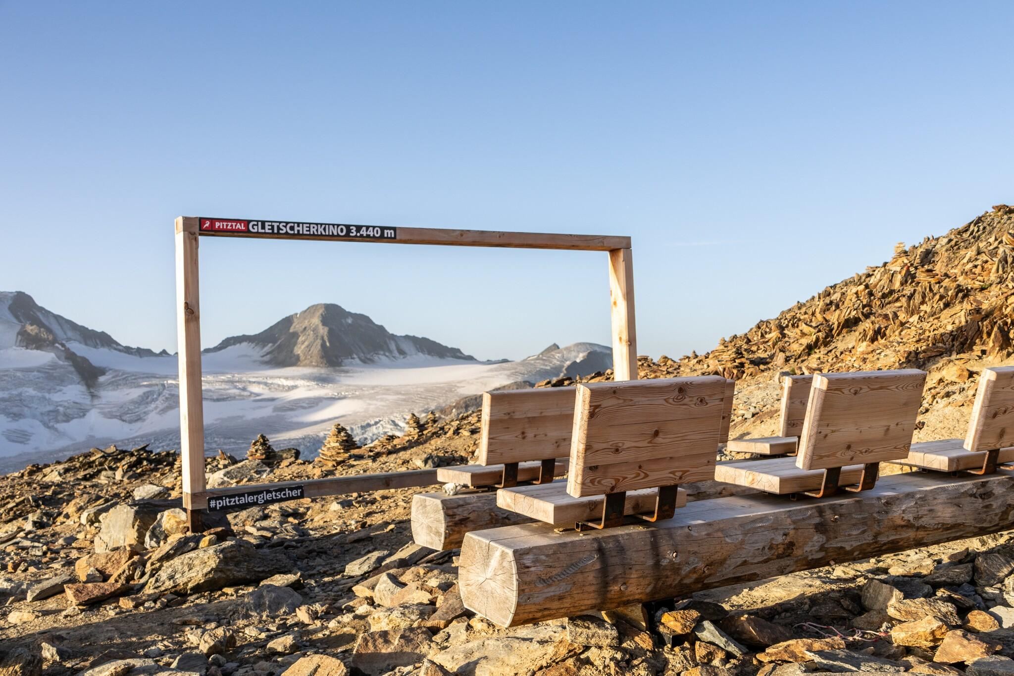 A picturesque view of the mountains with a wooden frame construction and benches in the foreground. The landscape features snow-capped peaks and a clear sky.