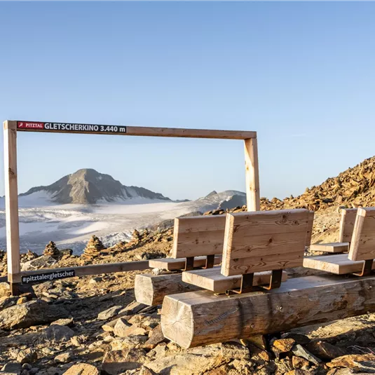 A picturesque view of the mountains with a wooden frame construction and benches in the foreground. The landscape features snow-capped peaks and a clear sky.