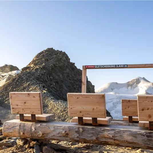 A wooden bench overlooking the mountains. In the background, snow-covered peaks and a clear sky can be seen.