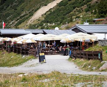 Ein gemütliches Restaurant in den Bergen mit Sonnenschirmen und Gästen auf der Terrasse. Die Umgebung ist grün und natürlich, umgeben von majestätischen Bergen.