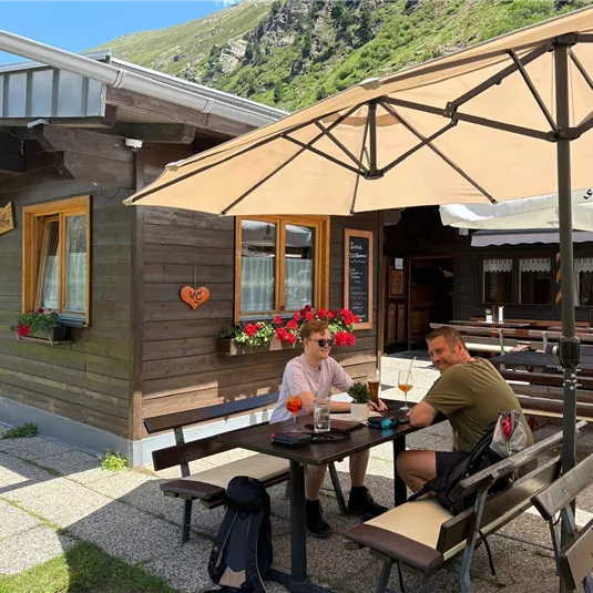 Eine gemütliche Außenansicht eines Holzrestaurants mit Tischen im Freien. Zwei Personen genießen Getränke und die Sonne unter einem Sonnenschirm.