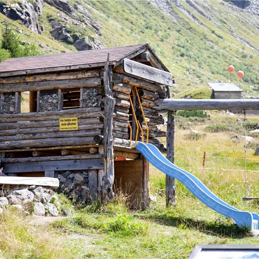 Ein kleines Spielhaus aus Holz mit einer Rutsche steht in einer grünen Wiesenlandschaft. Im Hintergrund sind Berge und farbige Ballons zu sehen.
