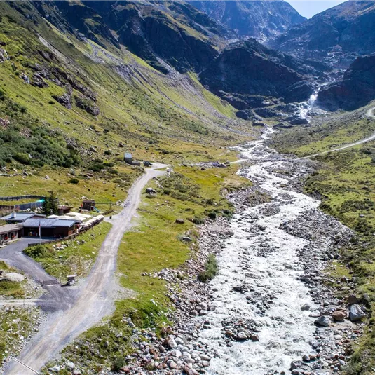 Eine malerische Talansicht mit einem klaren Fluss und grünen Wiesen. Im Hintergrund sind hohe Berge zu sehen.