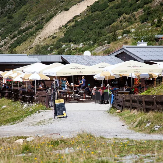 Ein gemütliches Restaurant in den Bergen mit sonnigen Terrassen und Sonnenschirmen. Viele Leute genießen die frische Luft und die schöne Umgebung.