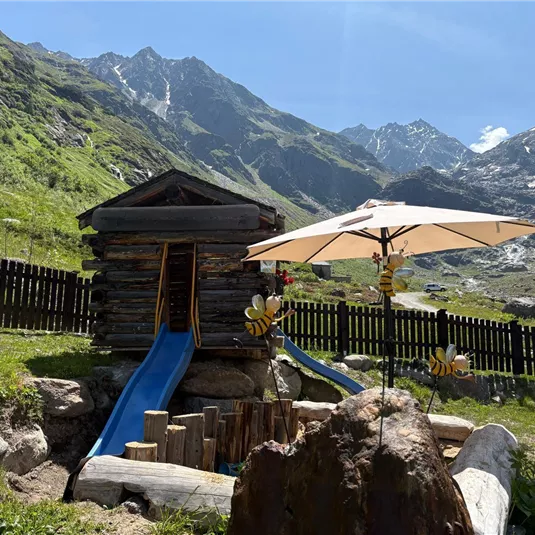 Ein Spielplatz mit einer Holzhütte und einer Rutsche, umgeben von grünen Wiesen und Bergen. Ein Sonnenschirm bietet Schatten auf dem Gelände.
