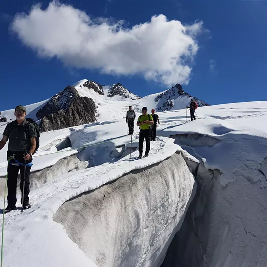Eine Gruppe von Menschen wandert über ein verschneites Gebirge. Im Hintergrund sind hohe Berge und ein blauer Himmel zu sehen.