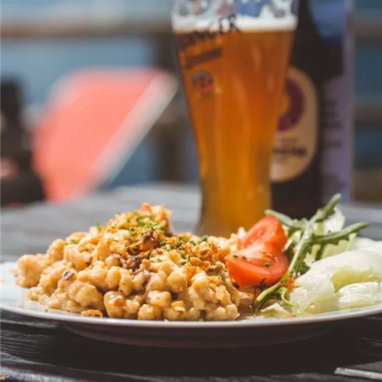 A plate with hearty food, consisting of pasta, garnished with herbs and fresh vegetables. In the background, there is a glass of beer.