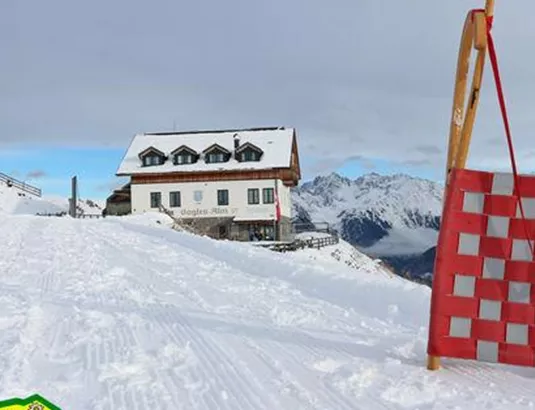 Eine verschneite Landschaft mit einem gemütlichen Berghaus im Hintergrund. Im Vordergrund steht ein Schlitten auf einer präparierten Piste.