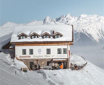 Eine schneebedeckte Berghütte umgeben von majestätischen Gipfeln. Die Szene zeigt eine ruhige Winterlandschaft mit klarem Himmel.