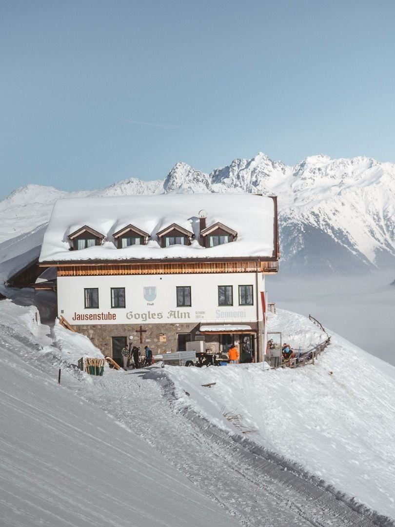 Eine schneebedeckte Berghütte umgeben von majestätischen Gipfeln. Die Szene zeigt eine ruhige Winterlandschaft mit klarem Himmel.