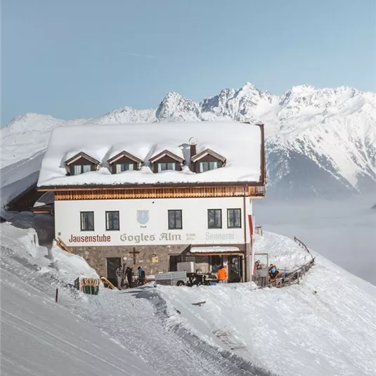 Eine schneebedeckte Berghütte umgeben von majestätischen Gipfeln. Die Szene zeigt eine ruhige Winterlandschaft mit klarem Himmel.