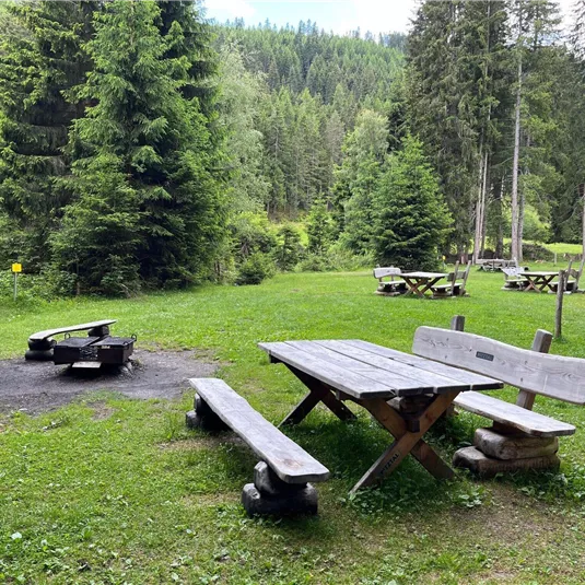 A cozy picnic spot in the forest with a wooden log table and benches. In the background, more seating and trees can be seen.
