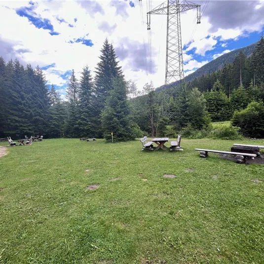Ein ruhiger, grüner Platz im Wald mit einigen Holzbänken und Tischen. Im Hintergrund sind hohe Bäume und ein blauer Himmel mit Wolken zu sehen.