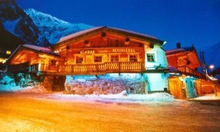 Ein gemütliches Chalet im winterlichen Bergdorf, beleuchtet in sanften Farben. Schnee bedeckt den Boden und die Berge sind im Hintergrund sichtbar.