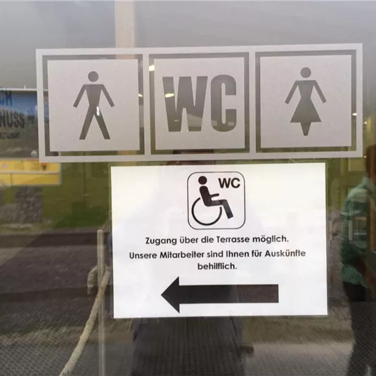 A sign with symbols for ladies, gentlemen, and accessible restroom. Below is information about access and assistance services.