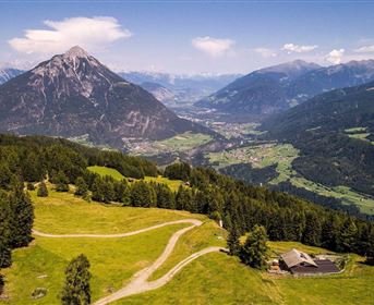 Eine malerische Berglandschaft mit grünen Wiesen und einem majestätischen Berg im Hintergrund. Im Tal erstrecken sich Dörfer und Wälder unter einem klaren blauen Himmel.