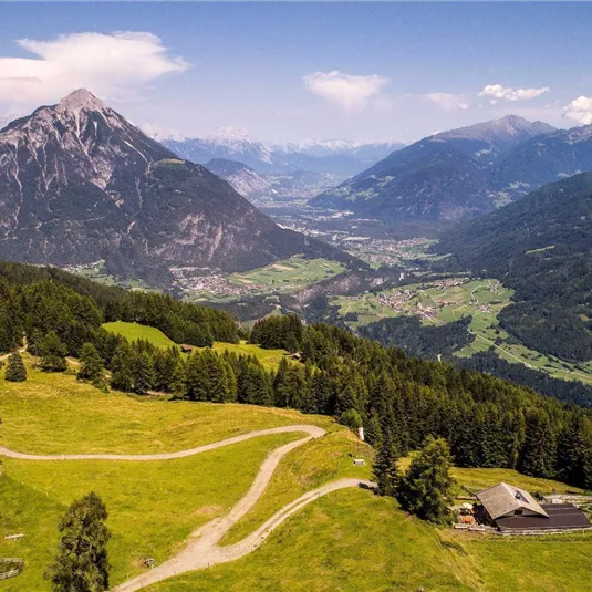 Eine malerische Berglandschaft mit grünen Wiesen und einem majestätischen Berg im Hintergrund. Im Tal erstrecken sich Dörfer und Wälder unter einem klaren blauen Himmel.
