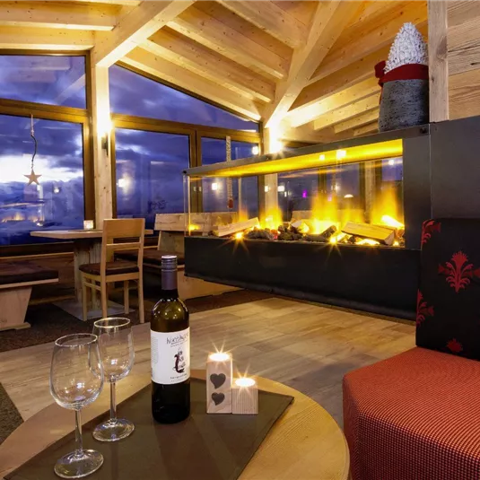 Ein gemütlicher Innenraum mit Holzelementen und einem Kamin. Auf dem Tisch stehen eine Flasche Wein und Gläser, was eine einladende Atmosphäre schafft.