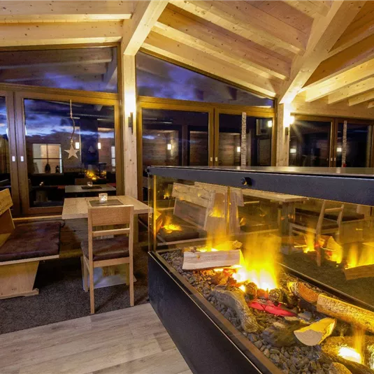 Ein gemütlicher Innenraum mit Holzmöbeln und einem modernen Kamin. Große Fenster bieten einen Blick auf den Abendhimmel.