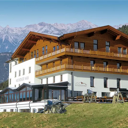 Ein modernes Hotelgebäude mit Holzverkleidung in einer bergigen Landschaft. Im Hintergrund sind hohe Berge und ein klarer Himmel zu sehen.
