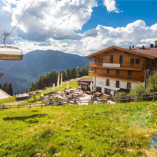 A restaurant in the mountains with a cable car next to it. The sky is blue with beautiful clouds and the surroundings are green and mountainous.
