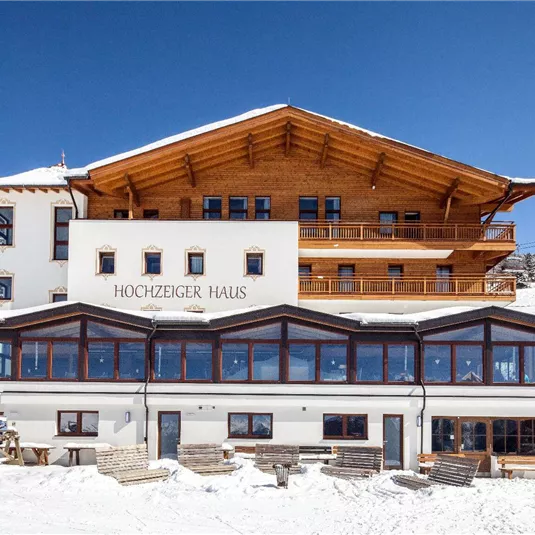 Ein alpines Hotel im Schnee mit Holzveranda und großen Fenstern. Im Hintergrund sind verschneite Bäume und ein klarer blauer Himmel zu sehen.