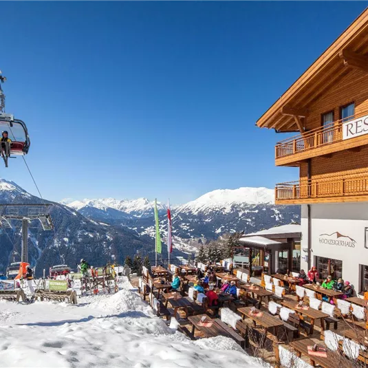 Eine Berglandschaft mit schneebedeckten Gipfeln und einem gemütlichen Restaurant. Im Vordergrund ist ein Skilift zu sehen, der Gäste befördert.