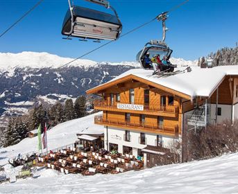 Ein gemütliches Restaurant in den Bergen, umgeben von Schnee. Eine Gondel fährt vorbei, während die Sonne die Landschaft erhellt.