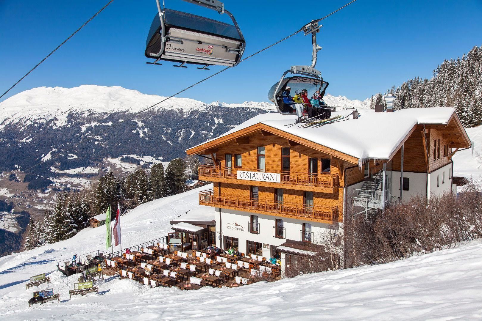 Ein gemütliches Restaurant in den Bergen, umgeben von Schnee. Eine Gondel fährt vorbei, während die Sonne die Landschaft erhellt.