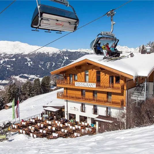 Ein gemütliches Restaurant in den Bergen, umgeben von Schnee. Eine Gondel fährt vorbei, während die Sonne die Landschaft erhellt.