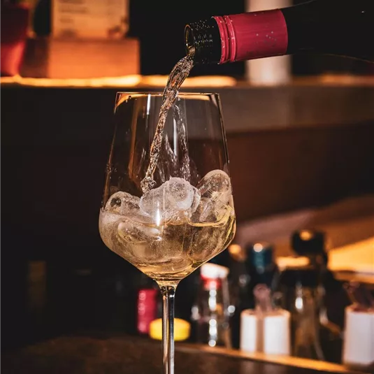 Ein Glas wird mit einem Getränk und Eiswürfeln gefüllt. Im Hintergrund sind verschiedene Flaschen sichtbar.