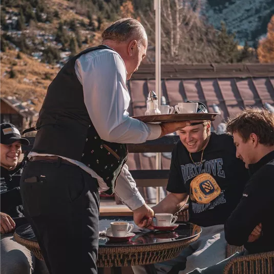 Eine gemütliche Szene in einem Bergrestaurant, wo ein Kellner Getränke an Gäste serviert. Im Hintergrund sind die Berge und der klare Himmel sichtbar.