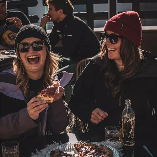 Zwei Frauen lachen und genießen Pizza am Tisch. Im Hintergrund sind andere Personen und eine helle Umgebung zu sehen.