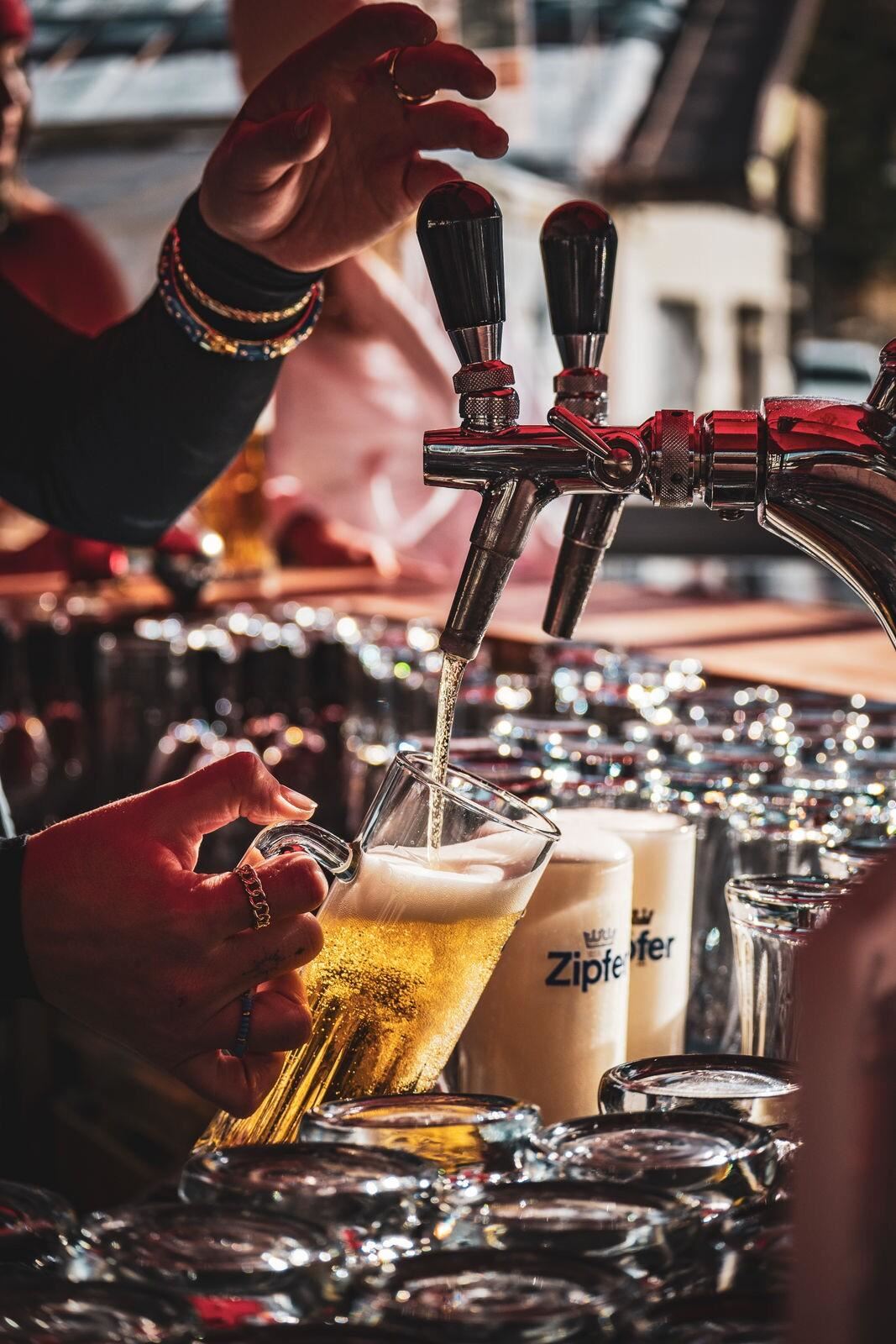 Ein Barmitarbeiter zapft ein kühles Bier in ein Glas. Im Hintergrund sind weitere Biergläser und Flaschen zu sehen.