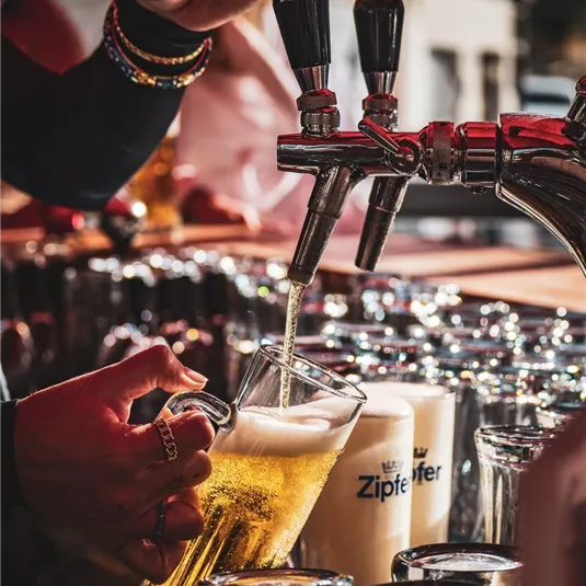 Ein Barmitarbeiter zapft ein kühles Bier in ein Glas. Im Hintergrund sind weitere Biergläser und Flaschen zu sehen.
