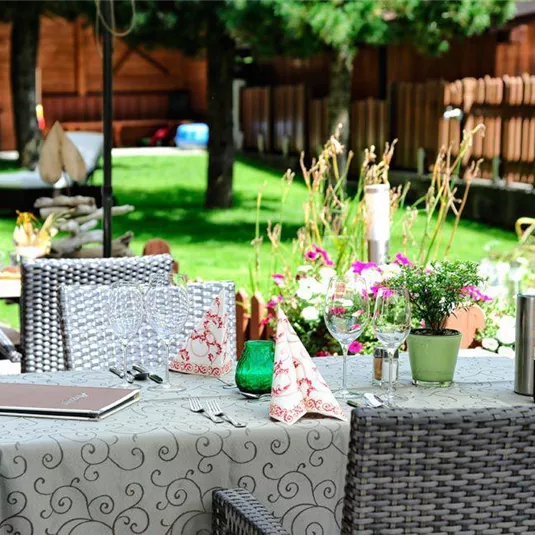 Ein einladender Tisch im Freien mit dekorativer Tischdecke, Gläsern und einer Flasche Wein. Im Hintergrund gibt es einen grünen Garten mit bunten Blumen und gemütlicher Atmosphäre.