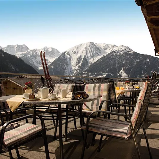 Eine gemütliche Terrasse mit Stühlen und Tischen, umgeben von Bergen und Schnee. Im Hintergrund strahlt ein klarer blauer Himmel.