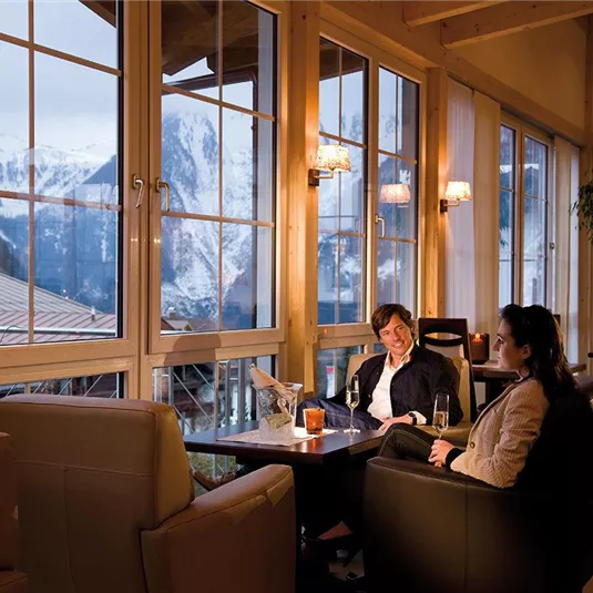 Eine gemütliche Lounge mit zwei Frauen, die an einem Tisch sitzen und Getränke genießen. Große Fenster bieten einen Blick auf die schneebedeckten Berge.